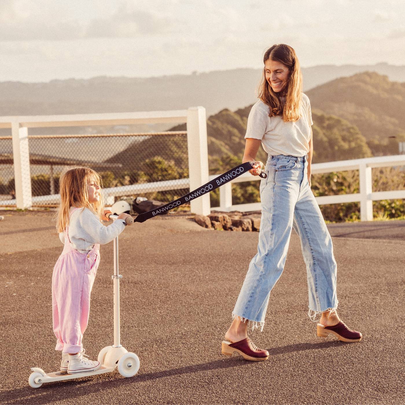 Woman holding black carry strap attached to child's scooter outdoors with sea backdrop