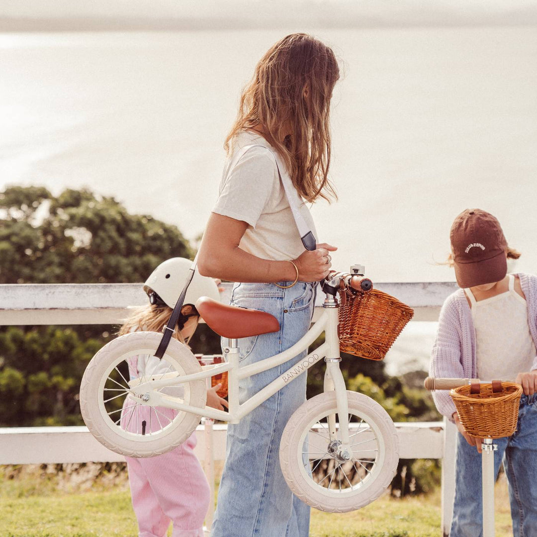 Girl carrying cream balance bike with brown basket using strap in outdoor countryside setting