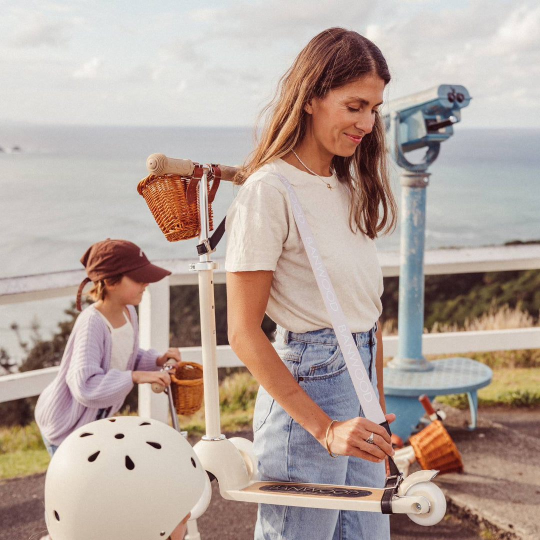 Girl carrying scooter with cream strap and basket in outdoor clifftop setting at dusk