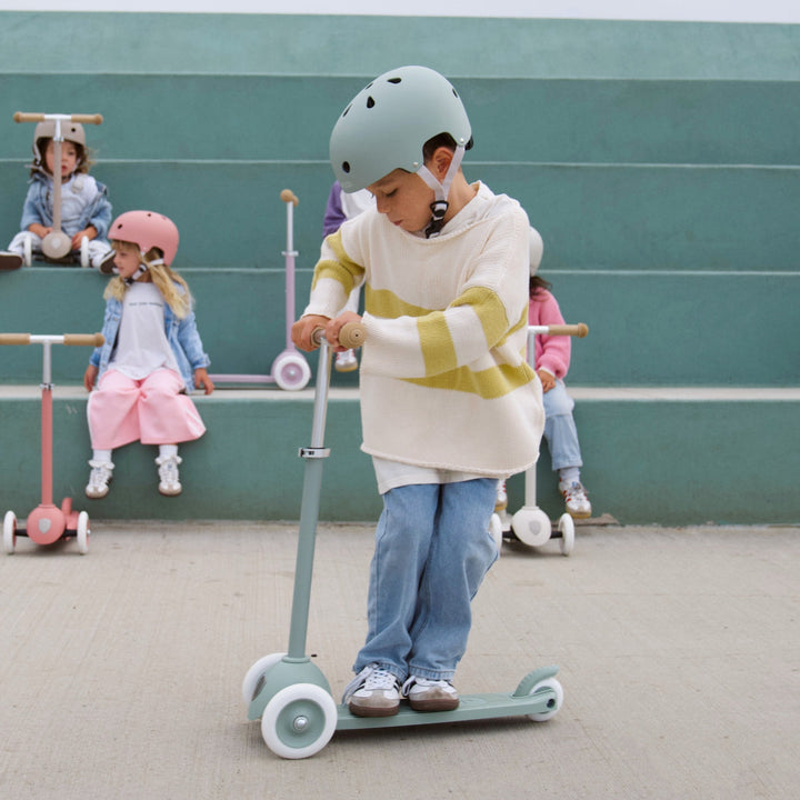 Child in sage Banwood helmet riding scooter outdoors with peers in background