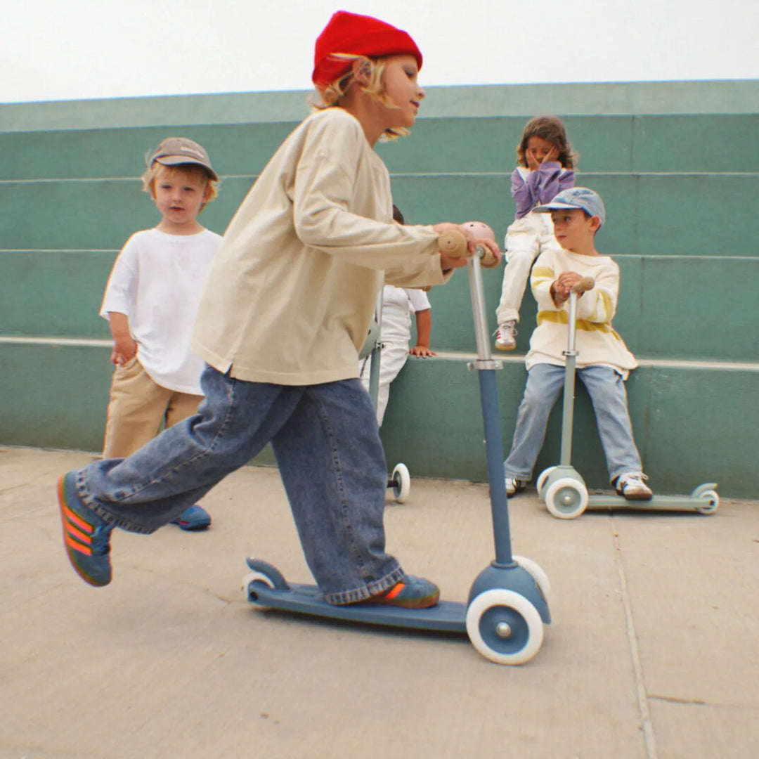 Child riding scooter on skate park with helmet and other young riders visible nearby