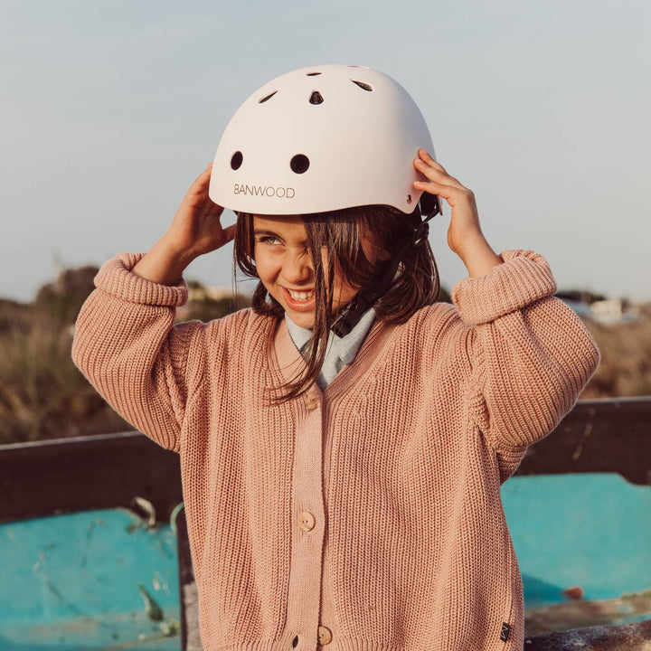 Child wearing pale pink helmet outdoors, hands to helmet, pink cardigan and seaside setting Banwood