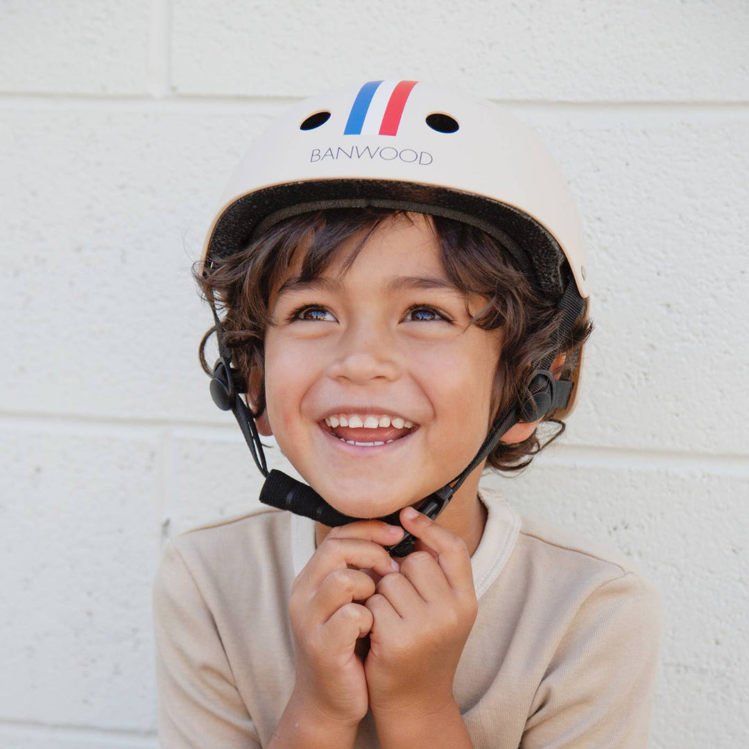 Child wearing cream Banwood striped helmet, securing chin strap with smile