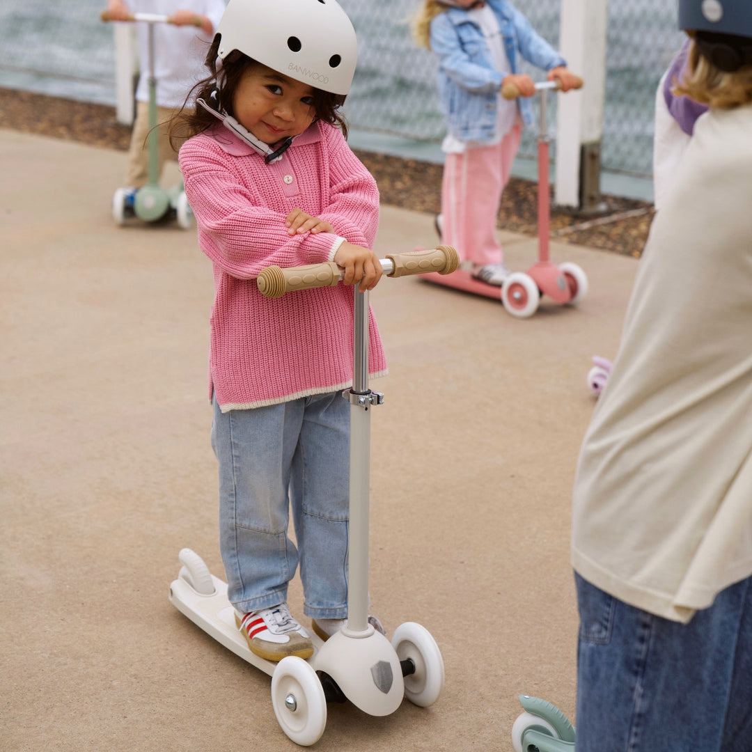 Child wearing cream Banwood helmet riding three-wheeled scooter at outdoor play area
