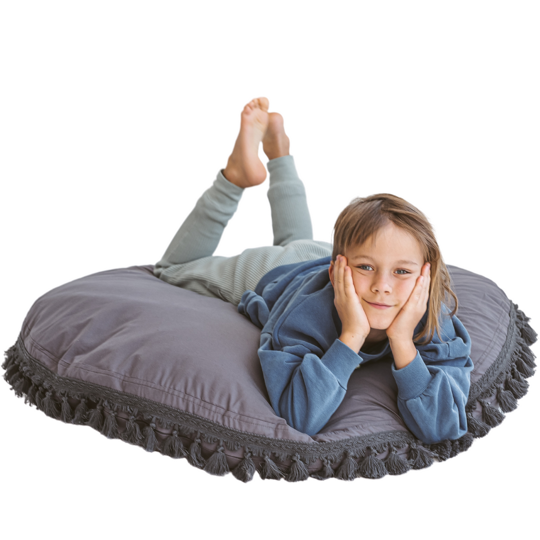 Child resting cheek on hands while lying on the MINICAMP grey floor cushion with tasselled trim, studio shot