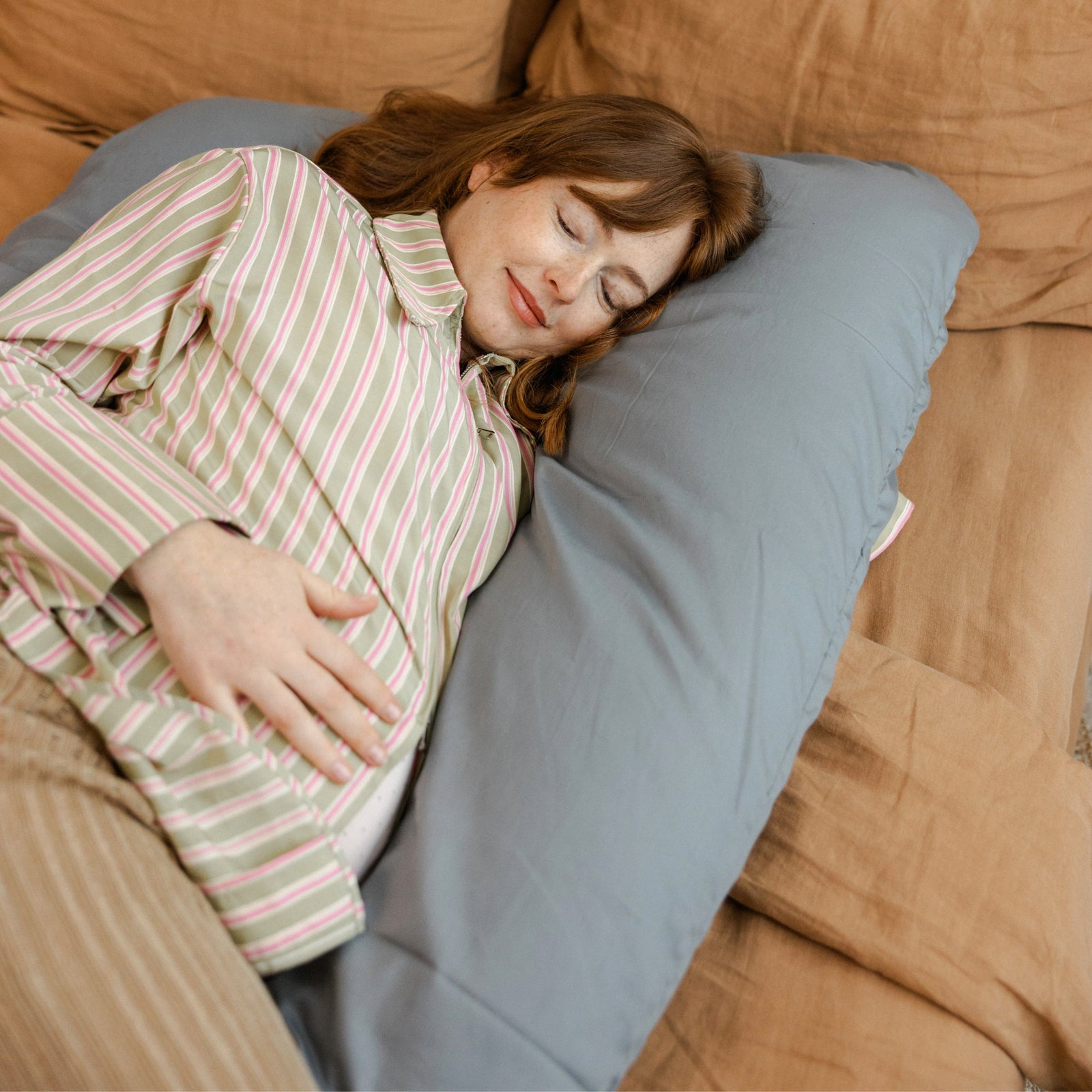 Pregnant woman resting with the MINICAMP U-shaped pregnancy pillow in stone grey, real bedroom setting