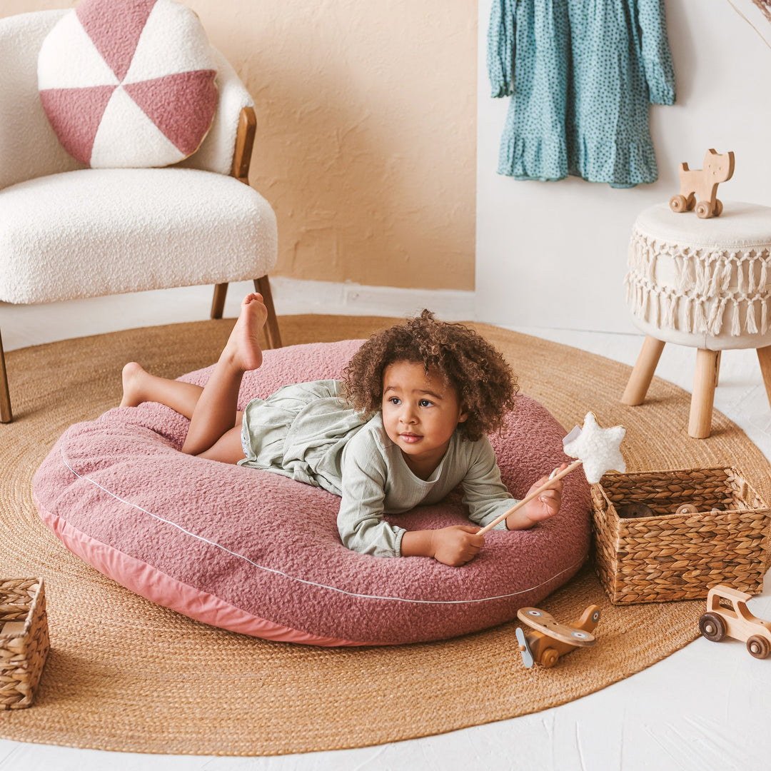 Child lying on the MINICAMP rose bouclé floor cushion in a boho-style nursery with a cream bouclé armchair and wicker baskets