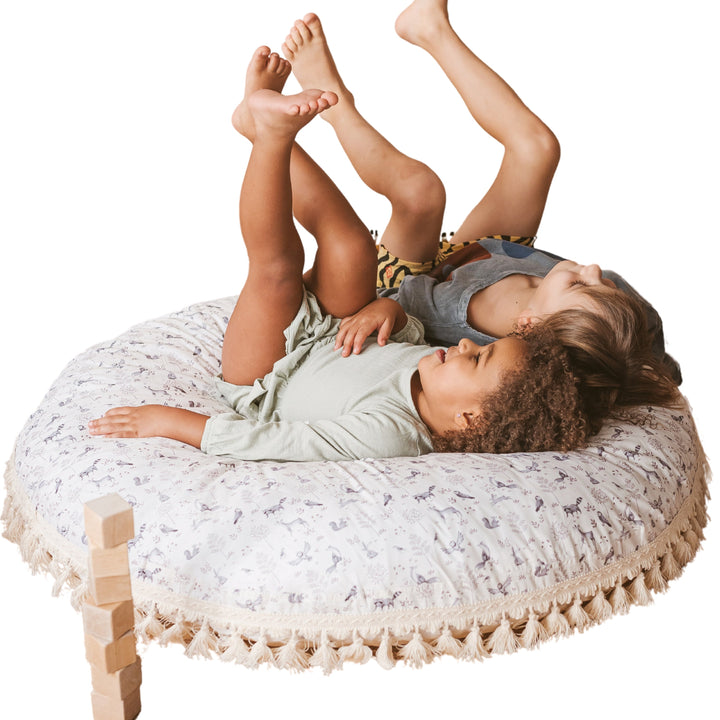 Two children lying on the MINICAMP forest animal print floor cushion with legs in the air, playful pose