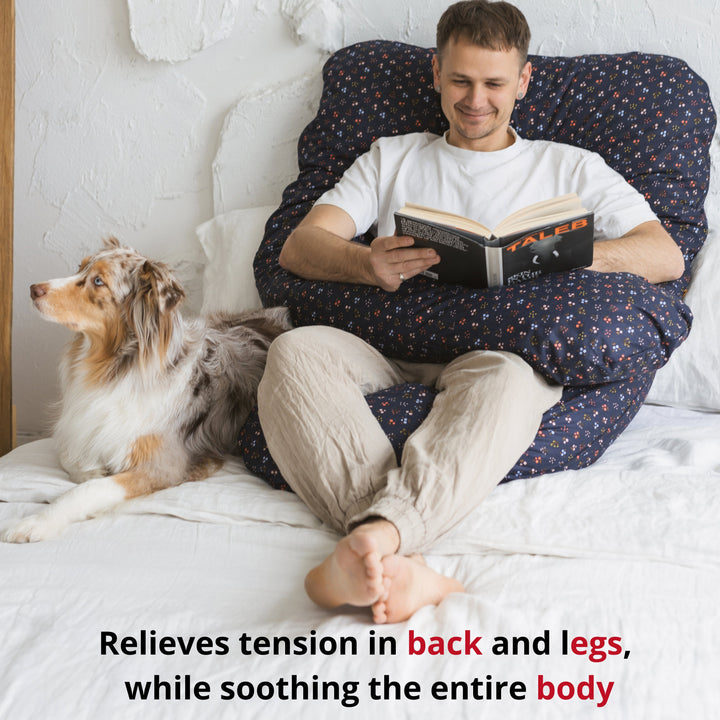 Man sitting up reading with a dog nearby, supported by the MINICAMP U-shaped comfort pillow in navy blue