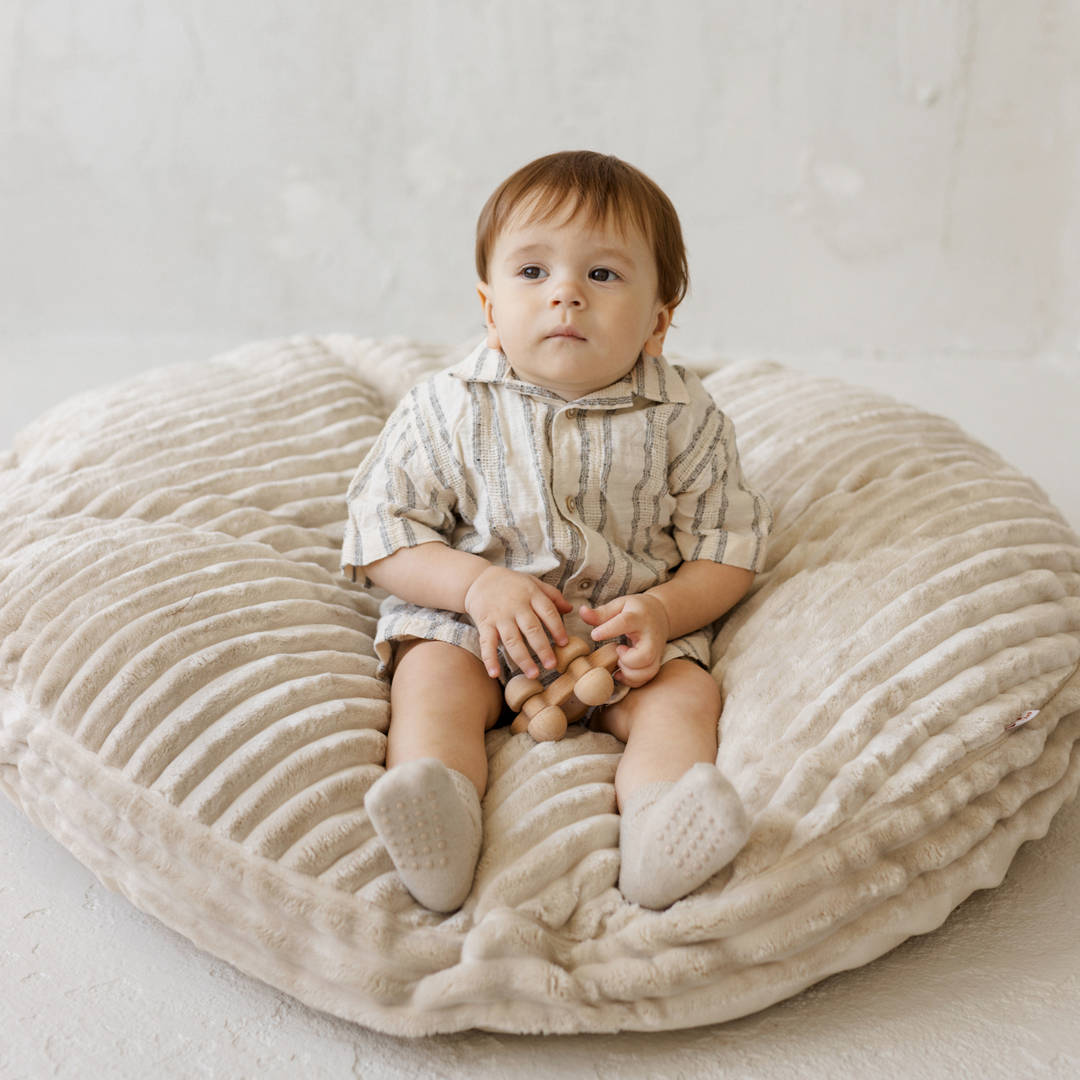 Baby sitting on the MINICAMP ecru corduroy floor cushion holding a wooden toy, front view