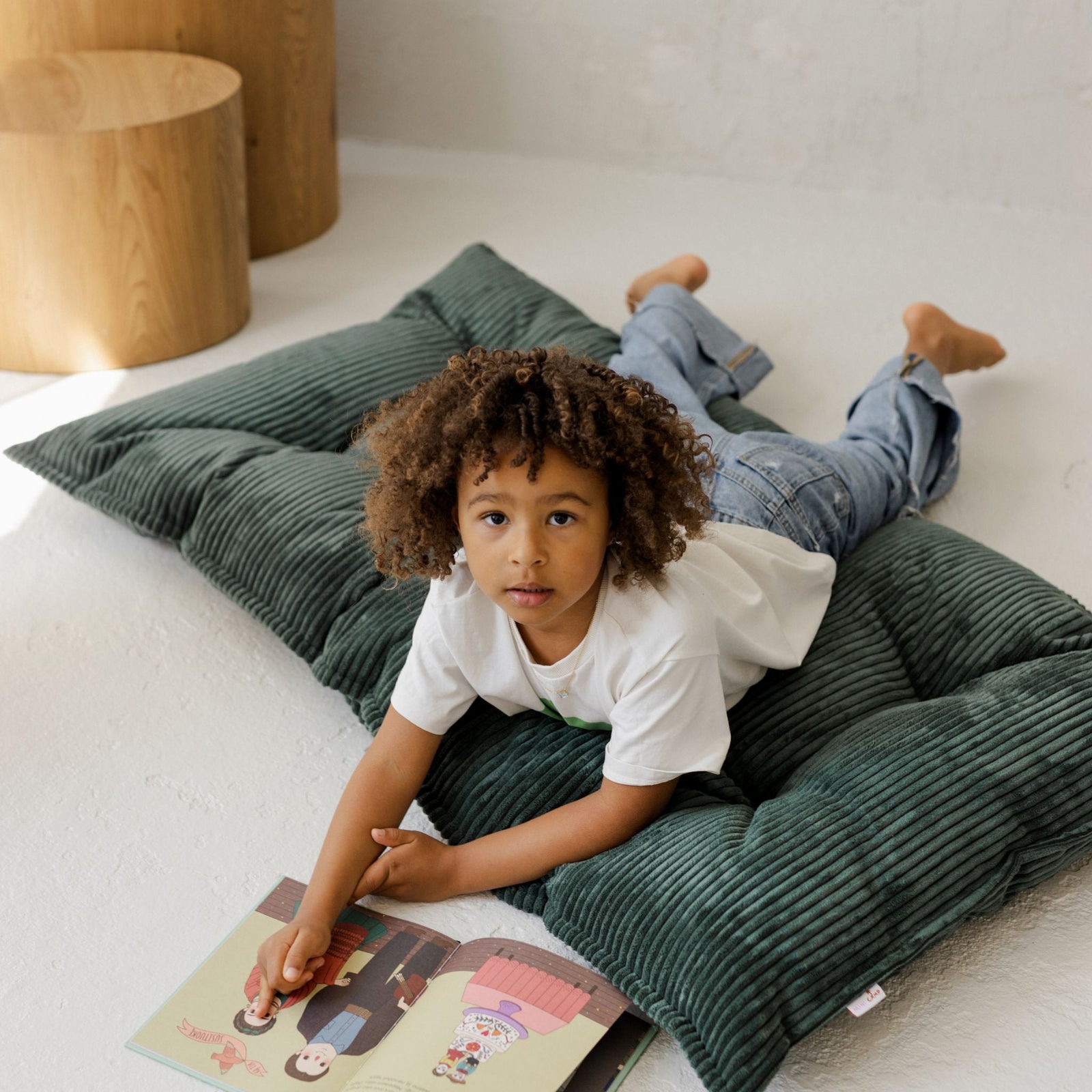 Child lying on the MINICAMP petrol green tufted floor cushion reading a book