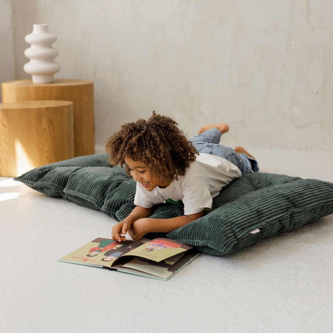 Child lying across the MINICAMP petrol green tufted floor cushion reading a book