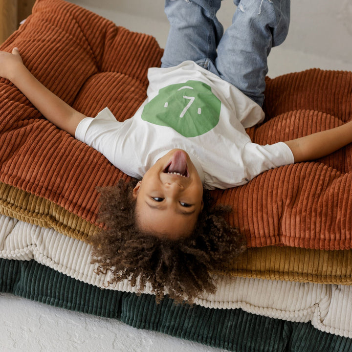 Child lying across the MINICAMP terracotta floor cushion stacked alongside petrol green, mustard and ecru colourways