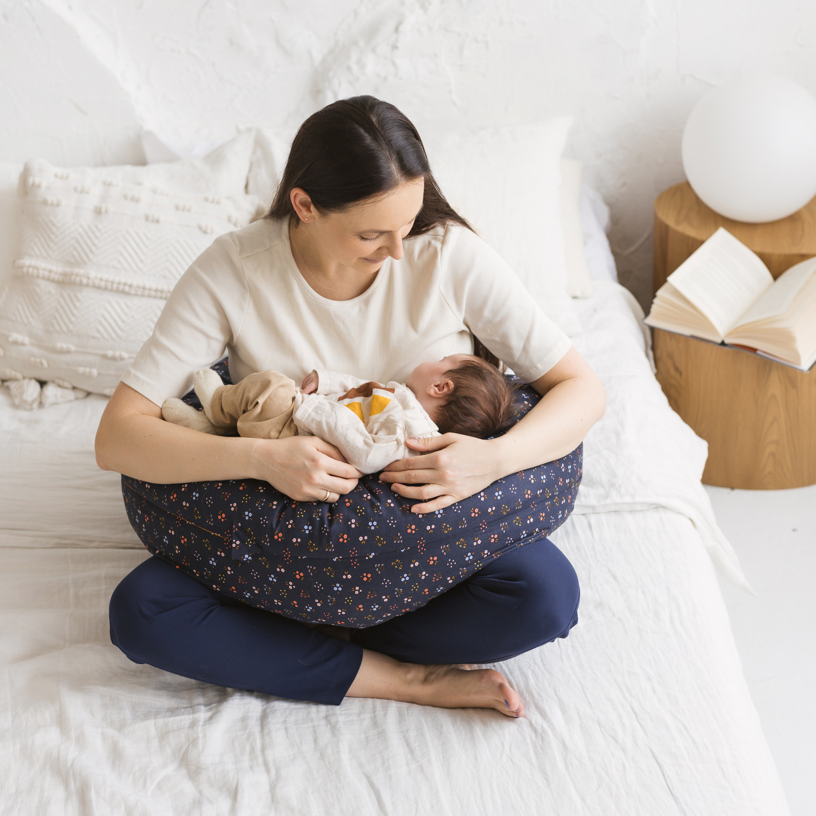 Woman sitting on a bed nursing her baby supported by the MINICAMP Navy Blue floral C-Shape Pillow