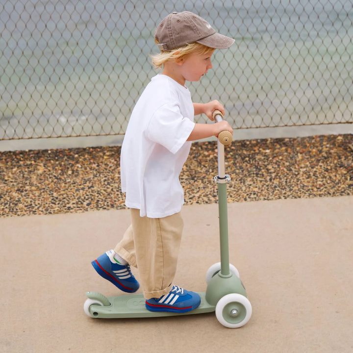 Child in white shirt and beige trousers standing on olive scooter against fence