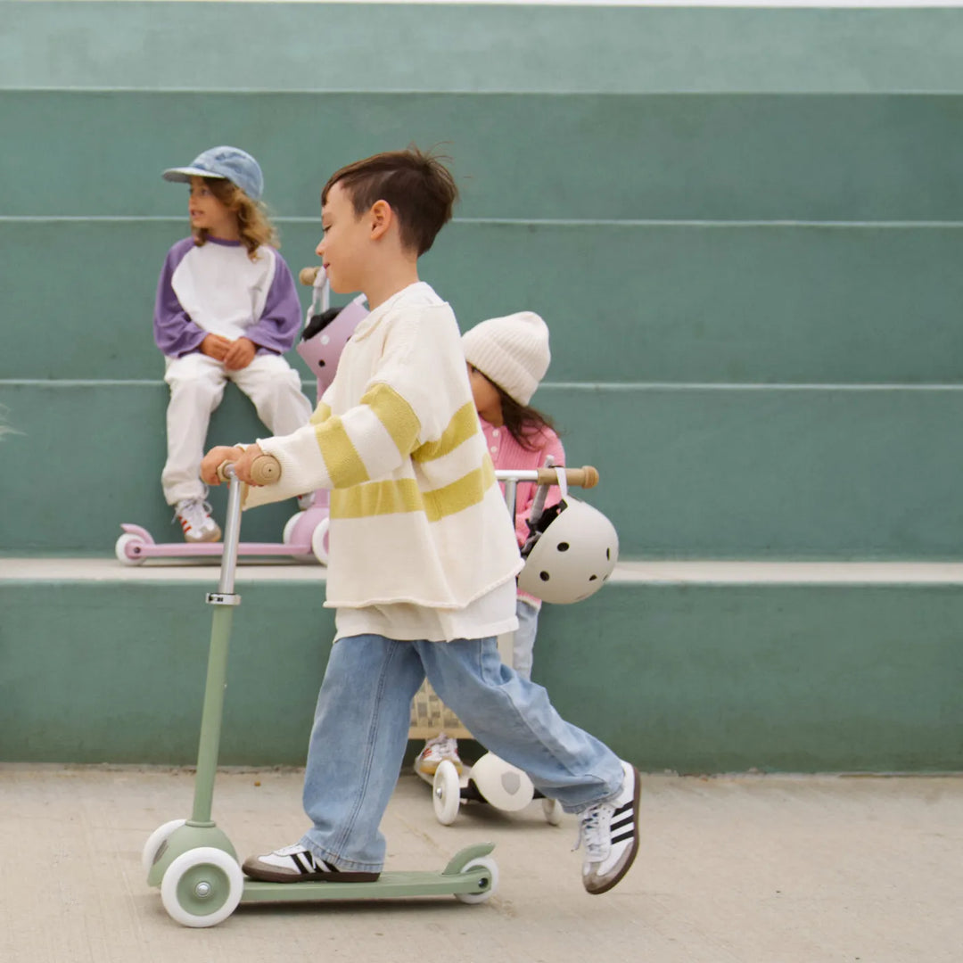 Three children riding scooters in pink, olive and purple colours against green wall