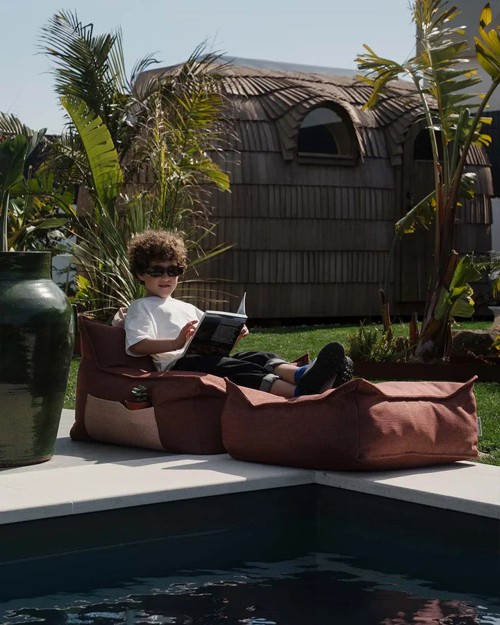 Child reading in burgundy bean bag chair beside pool, outdoor lifestyle