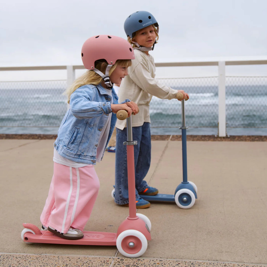 Two children on scooters wearing safety helmets, riding on beachside concrete with sea view
