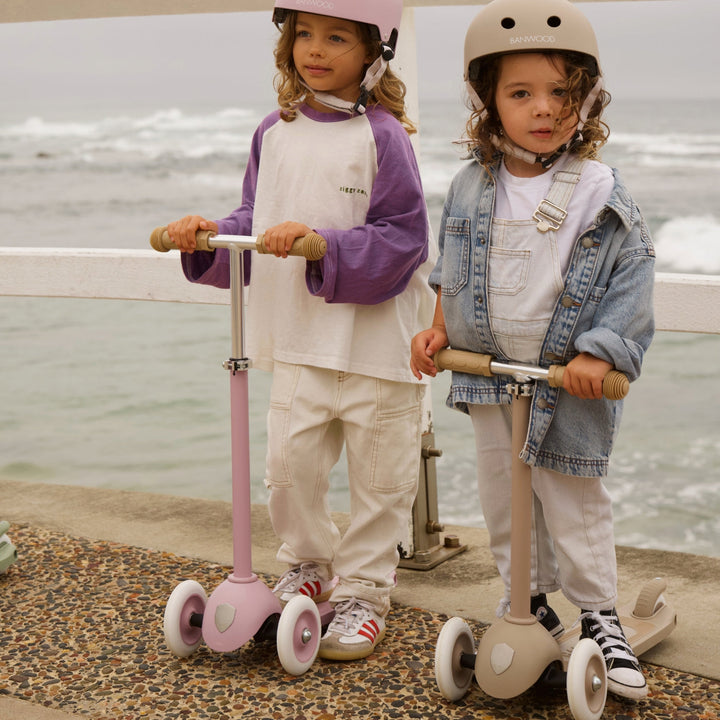 Two children wearing Banwood helmets riding scooters on sandy beach with sea behind