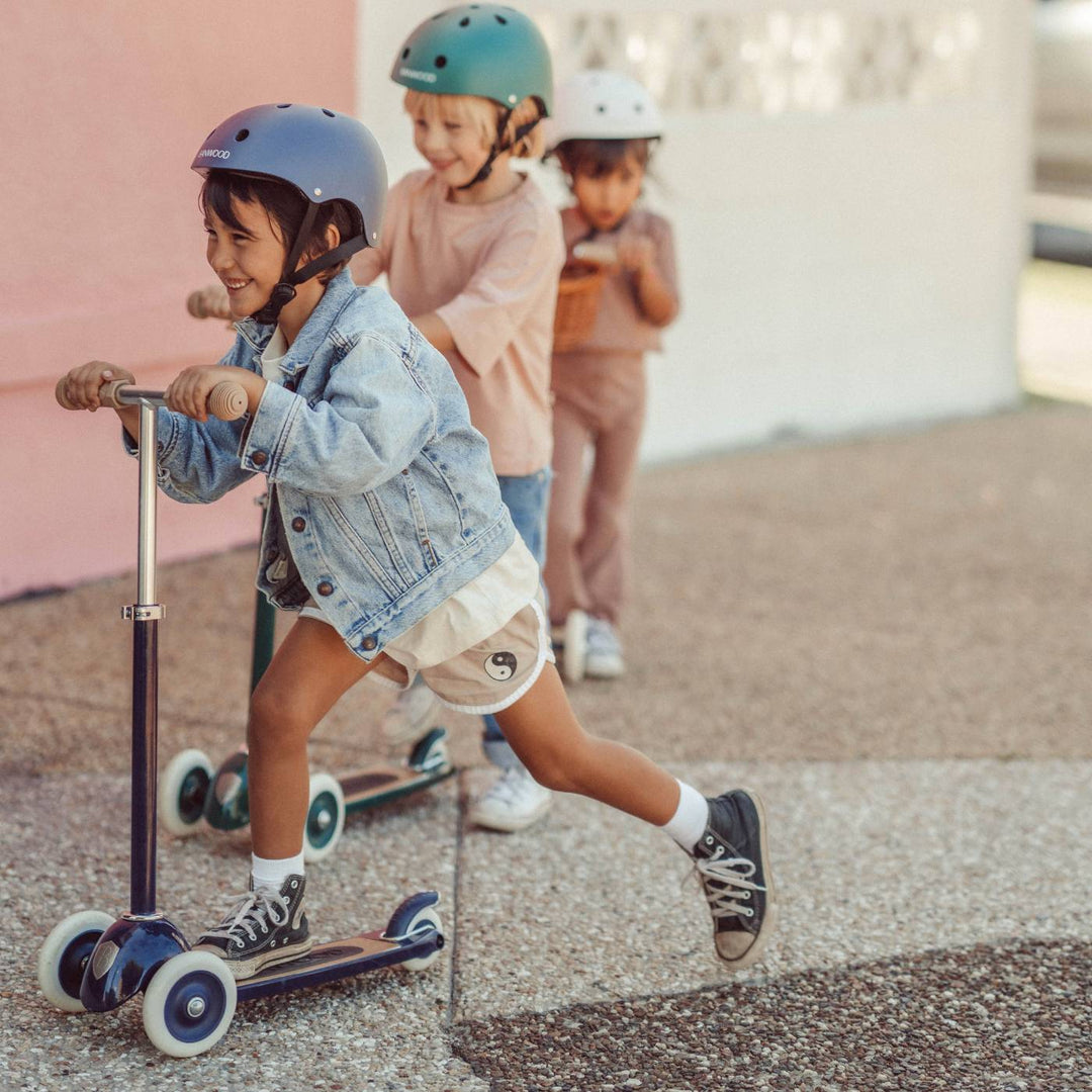 Child riding navy scooter with white wheels outdoors, two other children in background