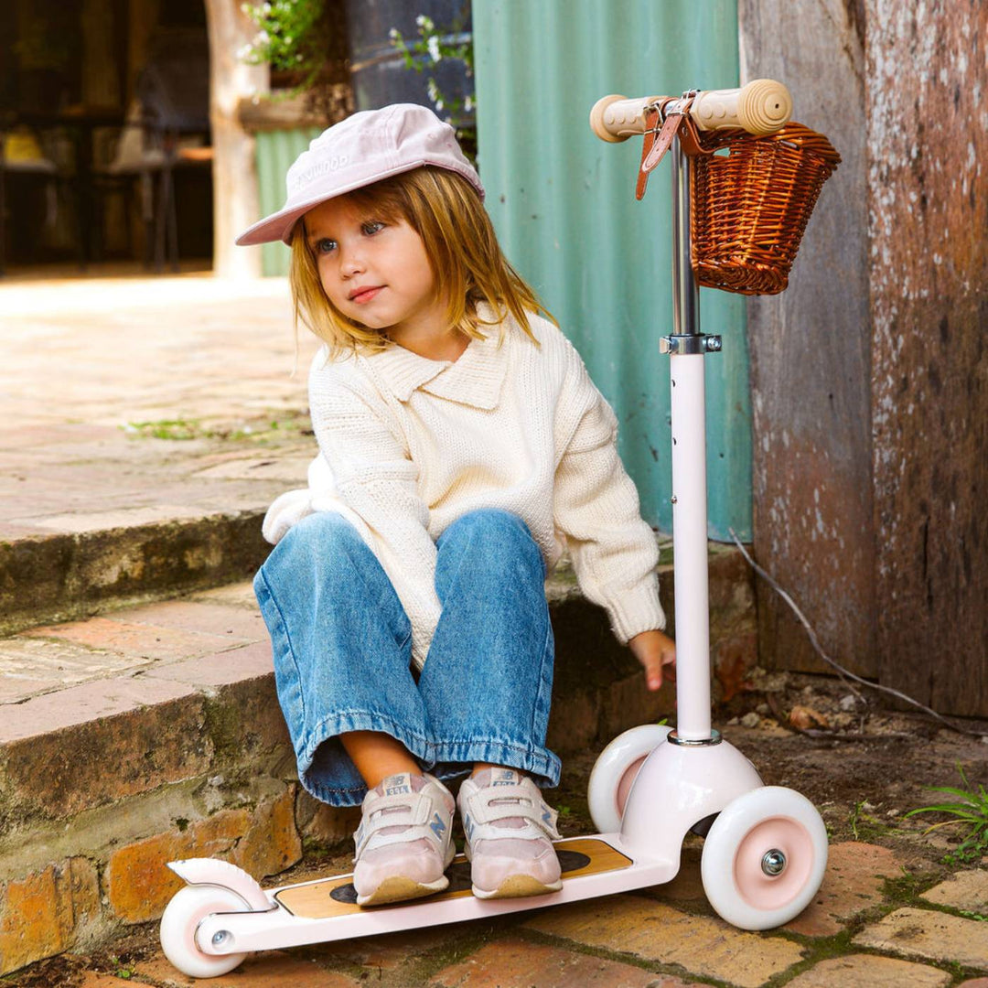 Child posing on pink scooter with wicker basket in outdoor urban setting