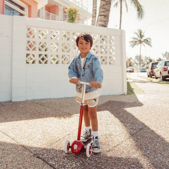 Child in denim jacket riding red Banwood scooter on driveway in daylight