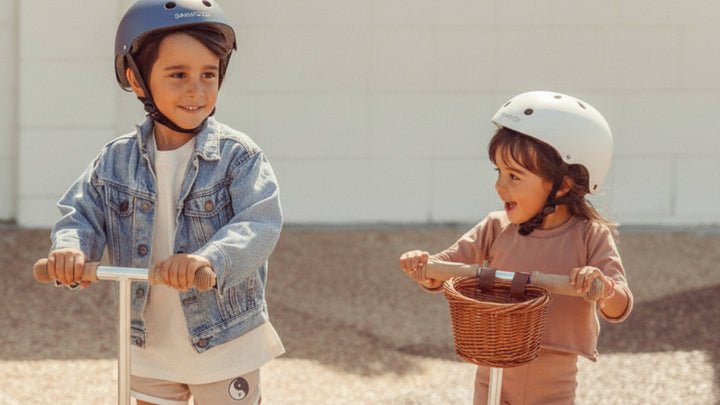 Two children riding white scooters with baskets on beach, wearing safety helmets