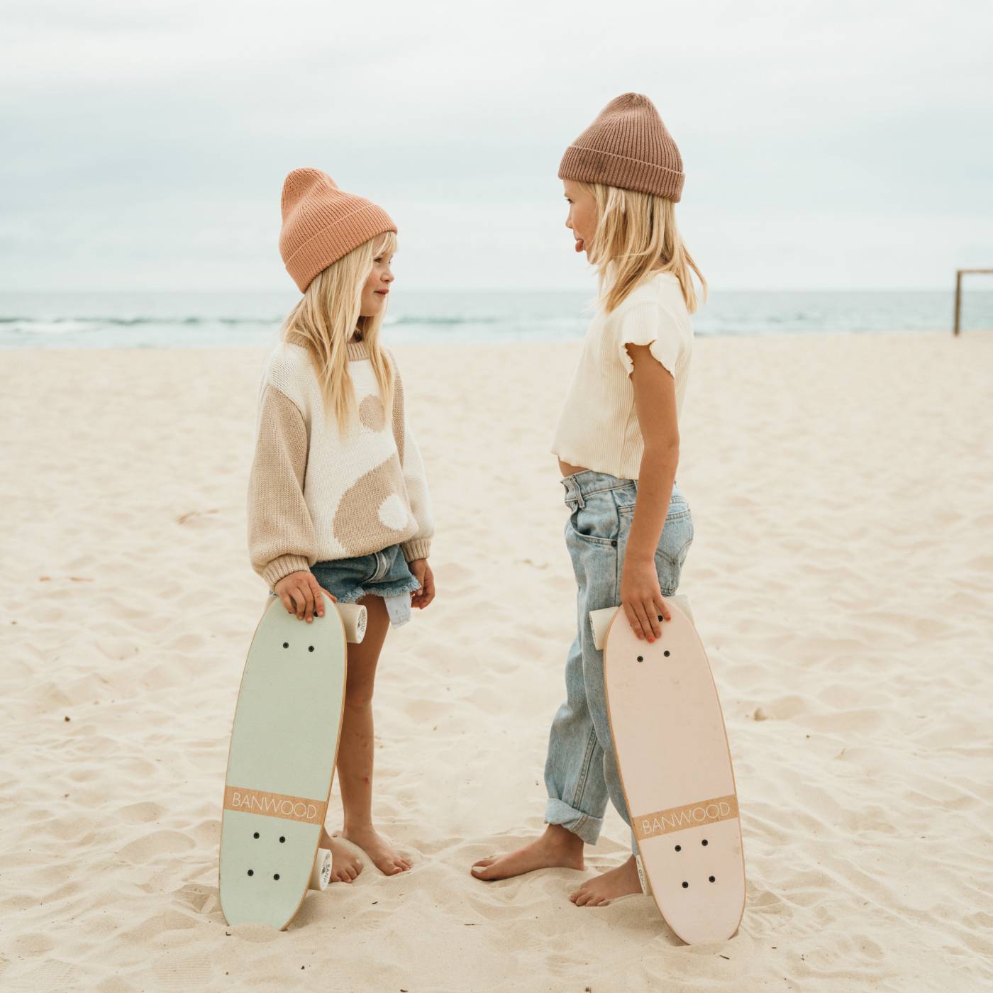 Two children on beach holding pink and mint skateboards, ocean background