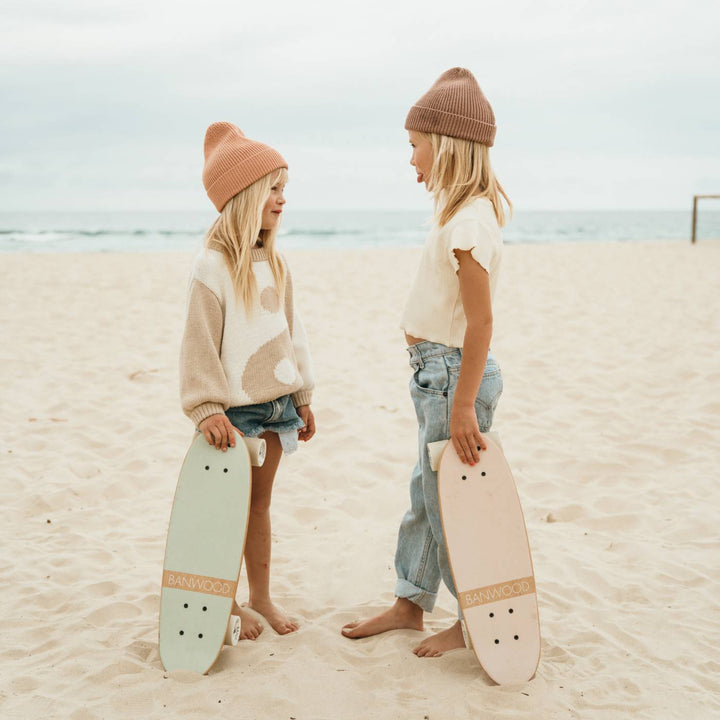 Two children on beach holding pink and mint skateboards, ocean background