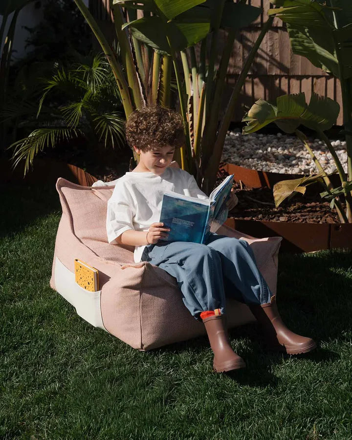 Child reclining in pink bean bag chair on grass, reading outdoors