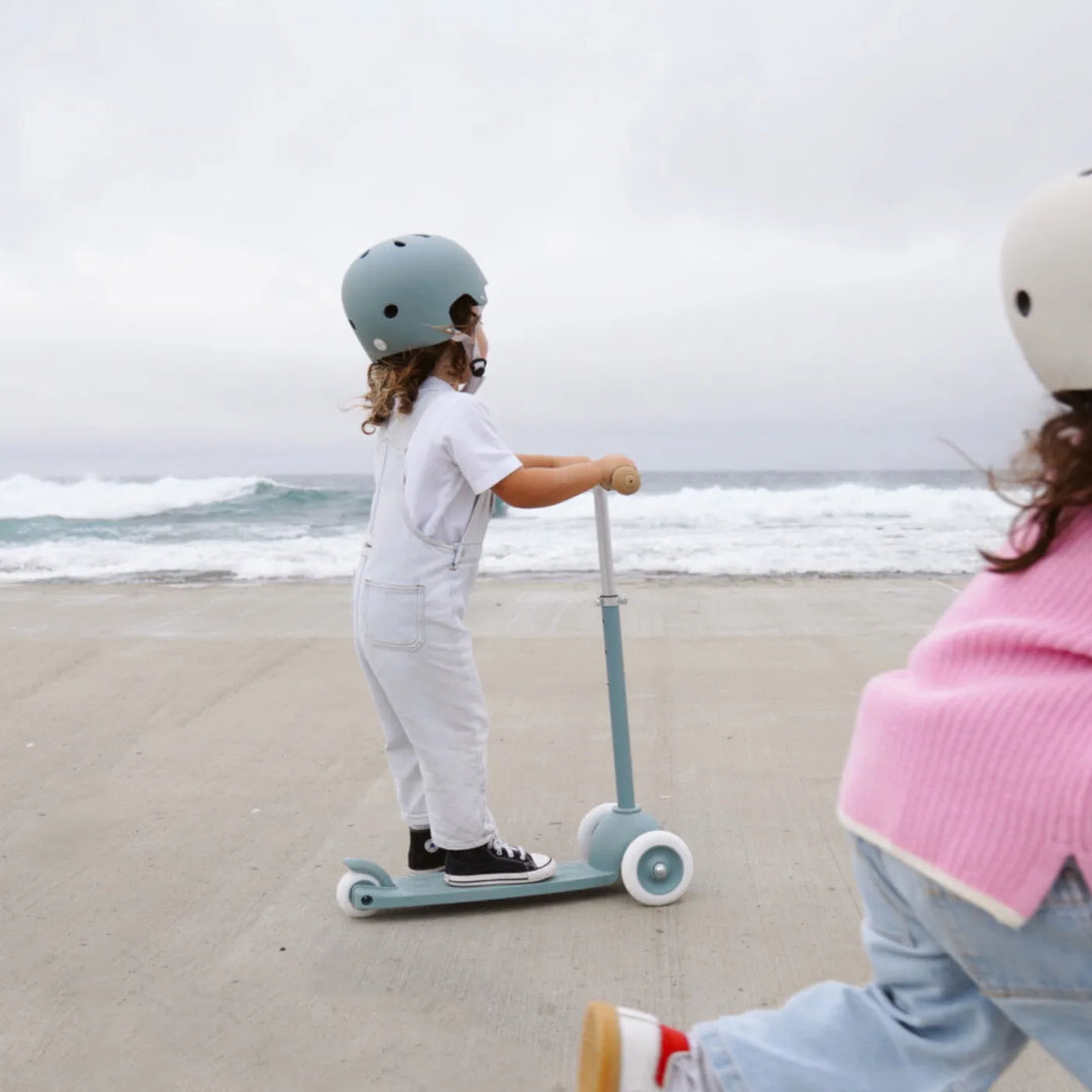 Child in teal helmet riding Banwood scooter on beach with sea and friend in background