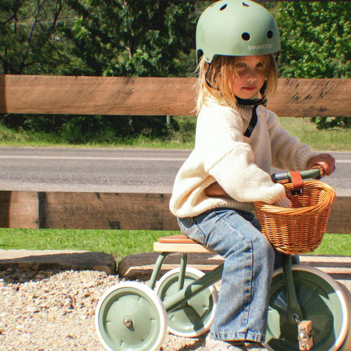Child in mint helmet riding sage green trike with wicker basket on garden path outdoors
