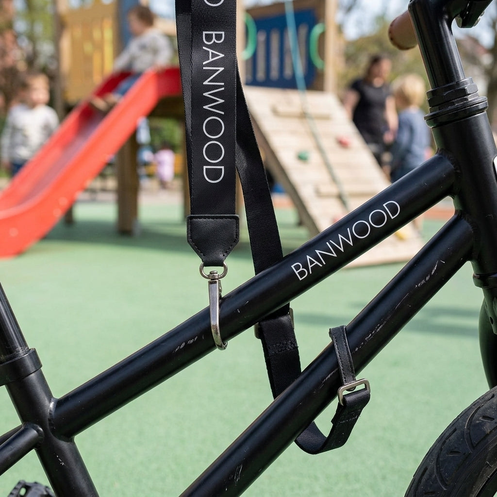 Close-up of the black Banwood carry strap clip fastened to a balance bike frame at a playground