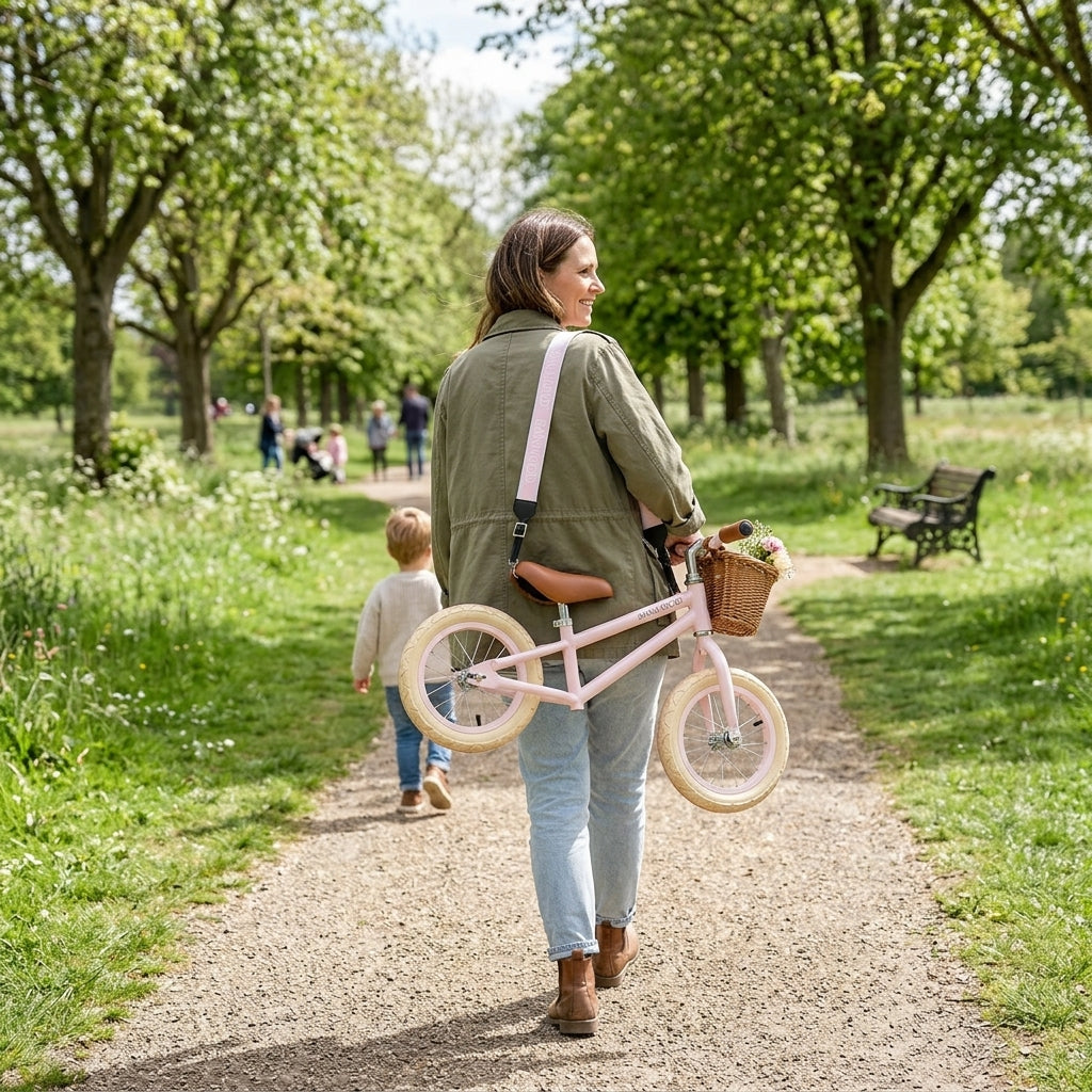 Parent carrying a pink Banwood balance bike over their shoulder using the Banwood carry strap while walking through a park