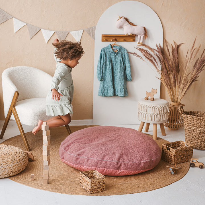 Child playing near the MINICAMP rose bouclé floor cushion in a decorated playroom with bunting and a unicorn plush toy
