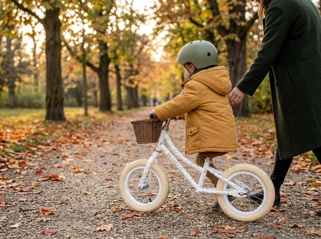 Child in a helmet on the Banwood Marest balance bike in Allegra White, an adult alongside on an autumn path