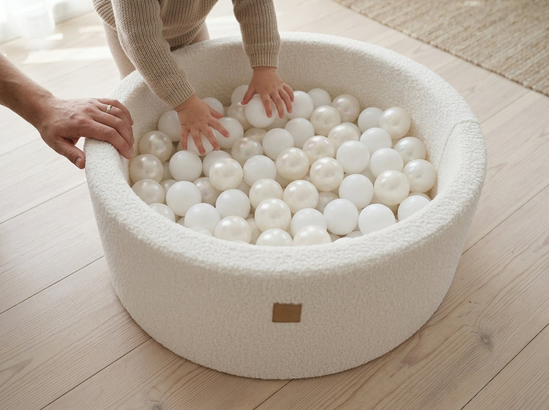 High-angle view of a toddler's hands reaching into the MeowBaby White Boucle Round Ball Pit, shown with play balls