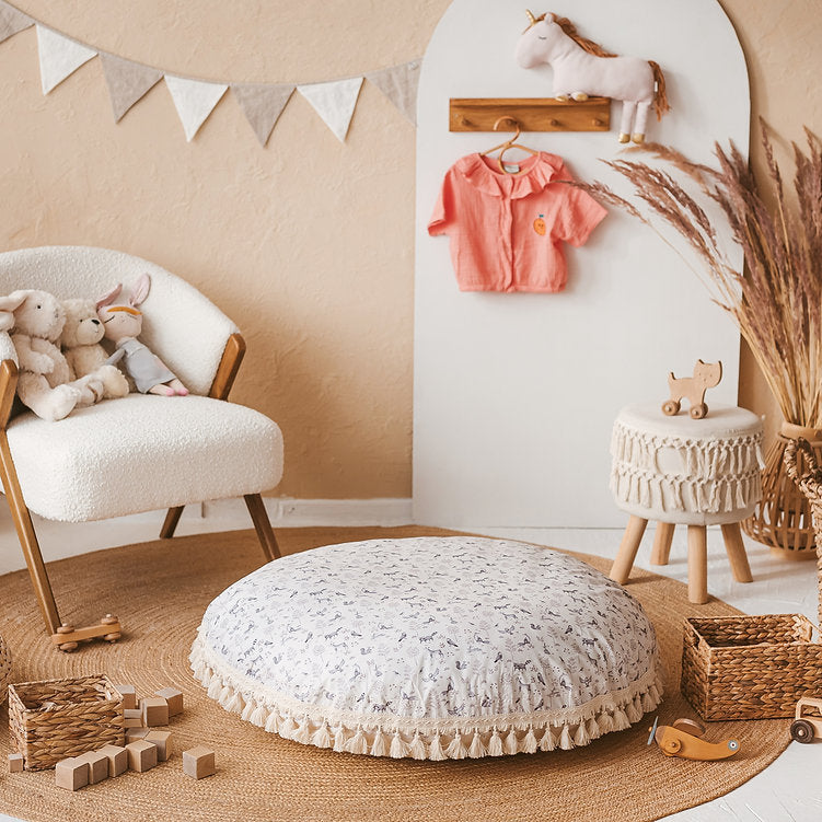 The MINICAMP forest animal print floor cushion with tassel trim, styled in a playroom with bunting and a plush unicorn