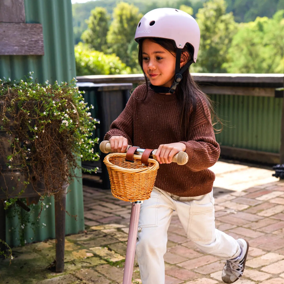 Child wearing pink Banwood helmet riding scooter on outdoor patio with basket