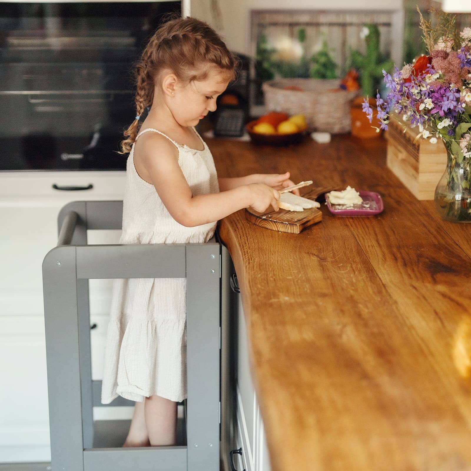 Child in grey kitchen helper tower at counter, using knife to chop food on board