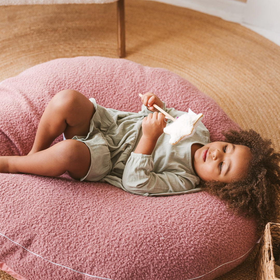 Close-up of a child lying on the MINICAMP rose bouclé floor cushion, holding a star wand toy
