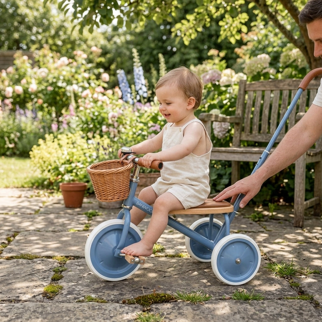 Toddler sitting on the Banwood Trike Vintage in Blue on a garden patio with a parent guiding the push handle