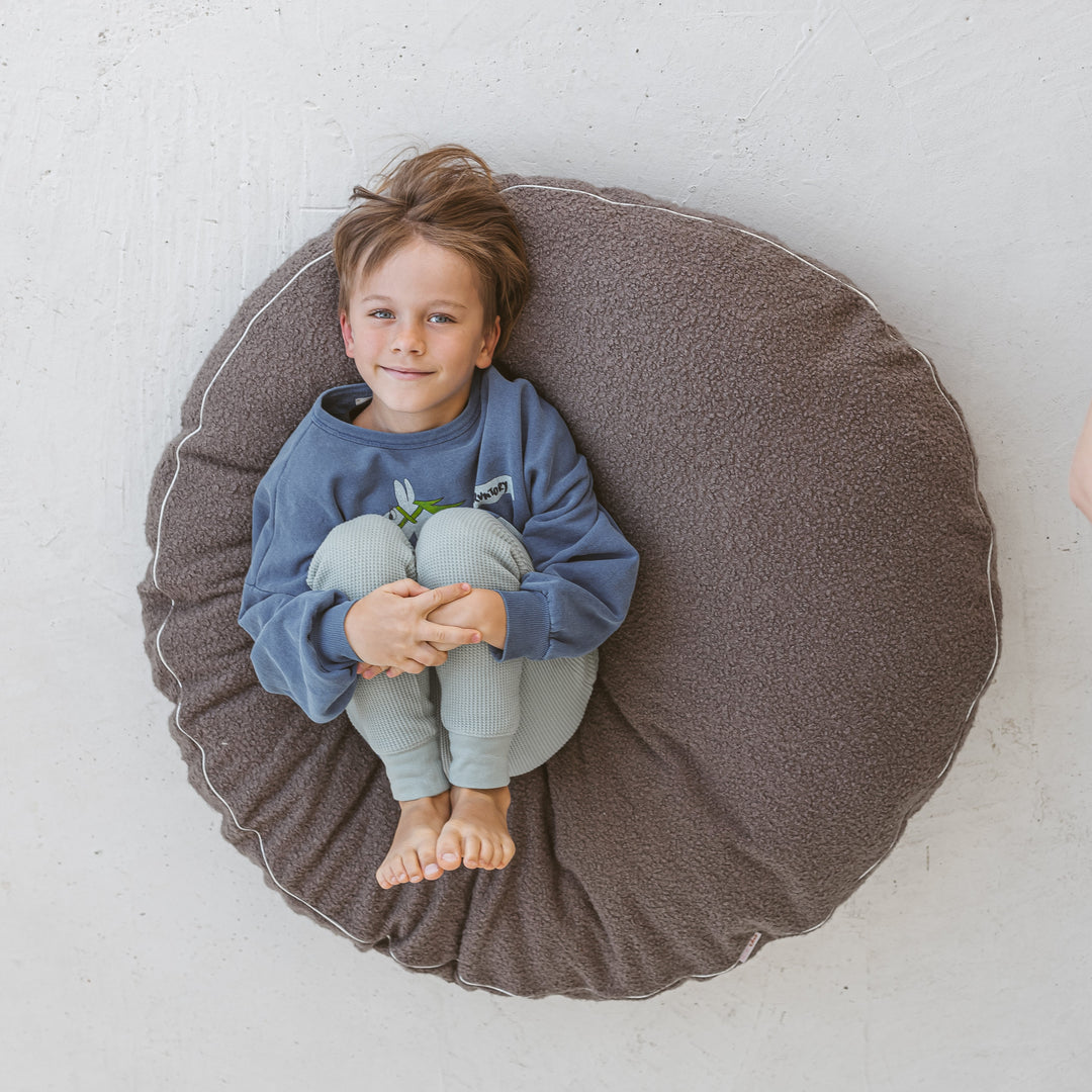 A boy sitting on the MINICAMP grey boucle floor cushion in a reading nook