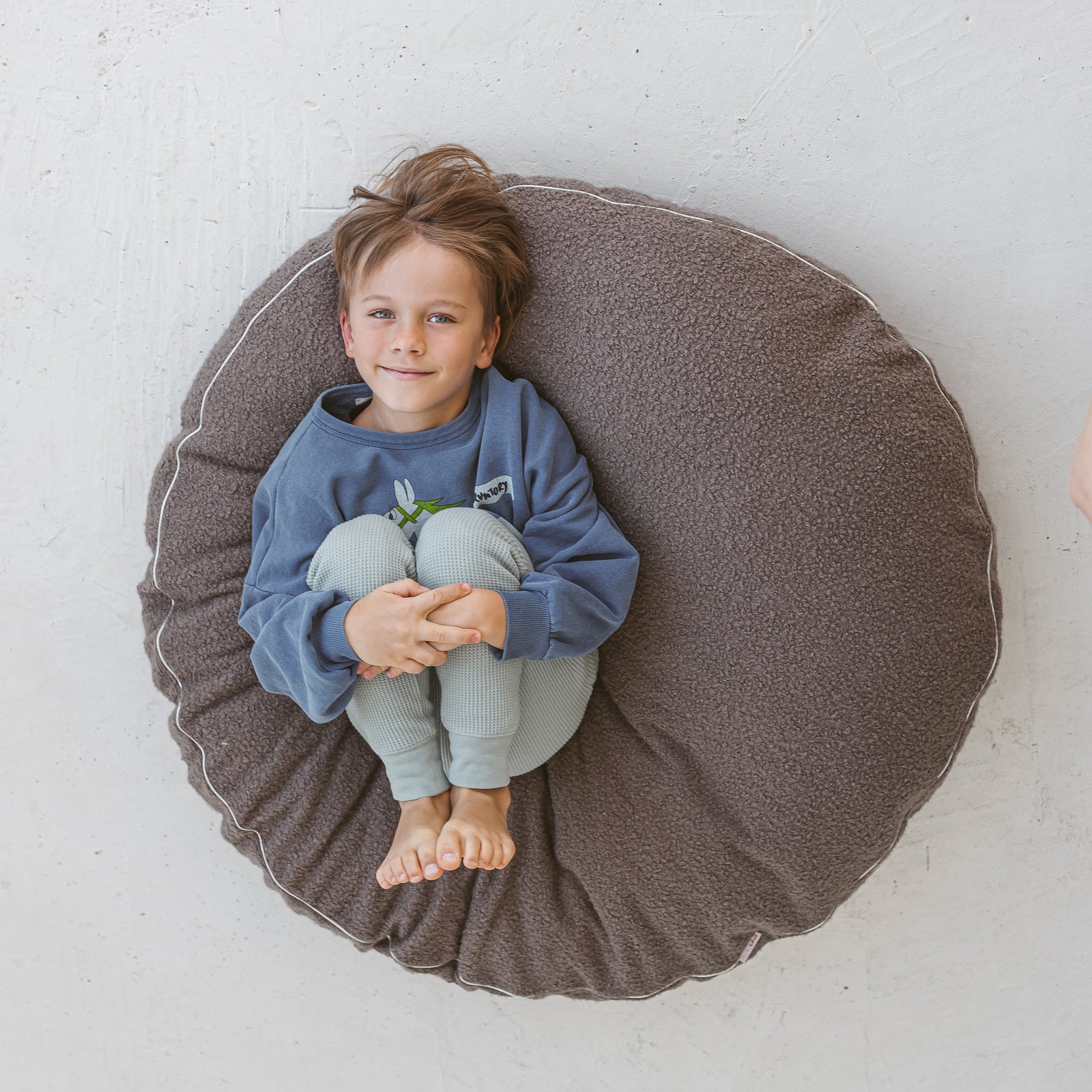 A boy sitting on the MINICAMP grey boucle floor cushion in a reading nook