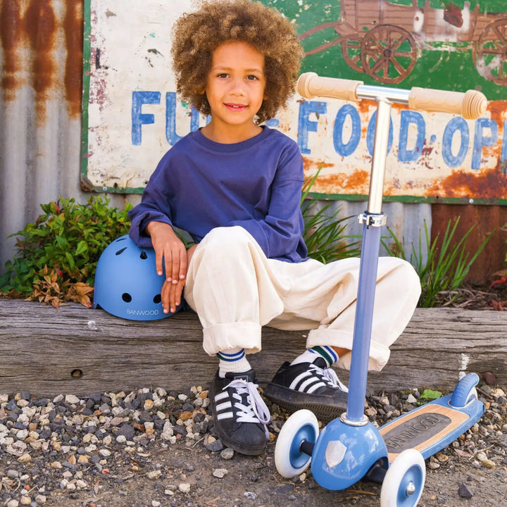 Child riding blue Banwood scooter outdoors, holding protective helmet