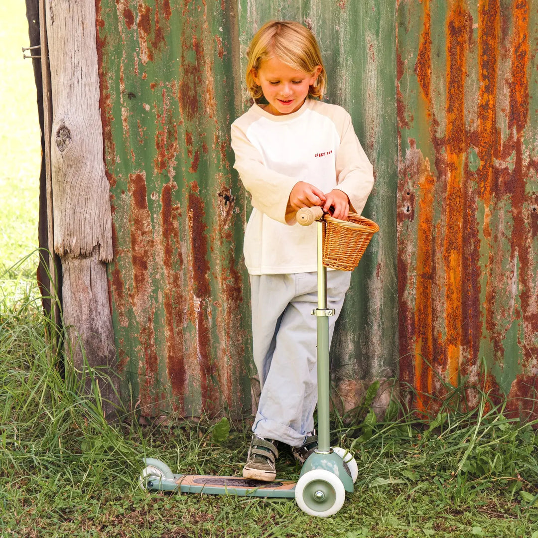 Child riding forest green scooter outdoors against rustic metal wall, wicker basket detail