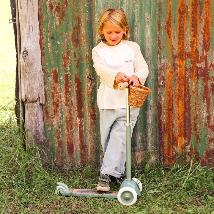 Child riding forest green scooter outdoors against rustic metal wall, wicker basket detail
