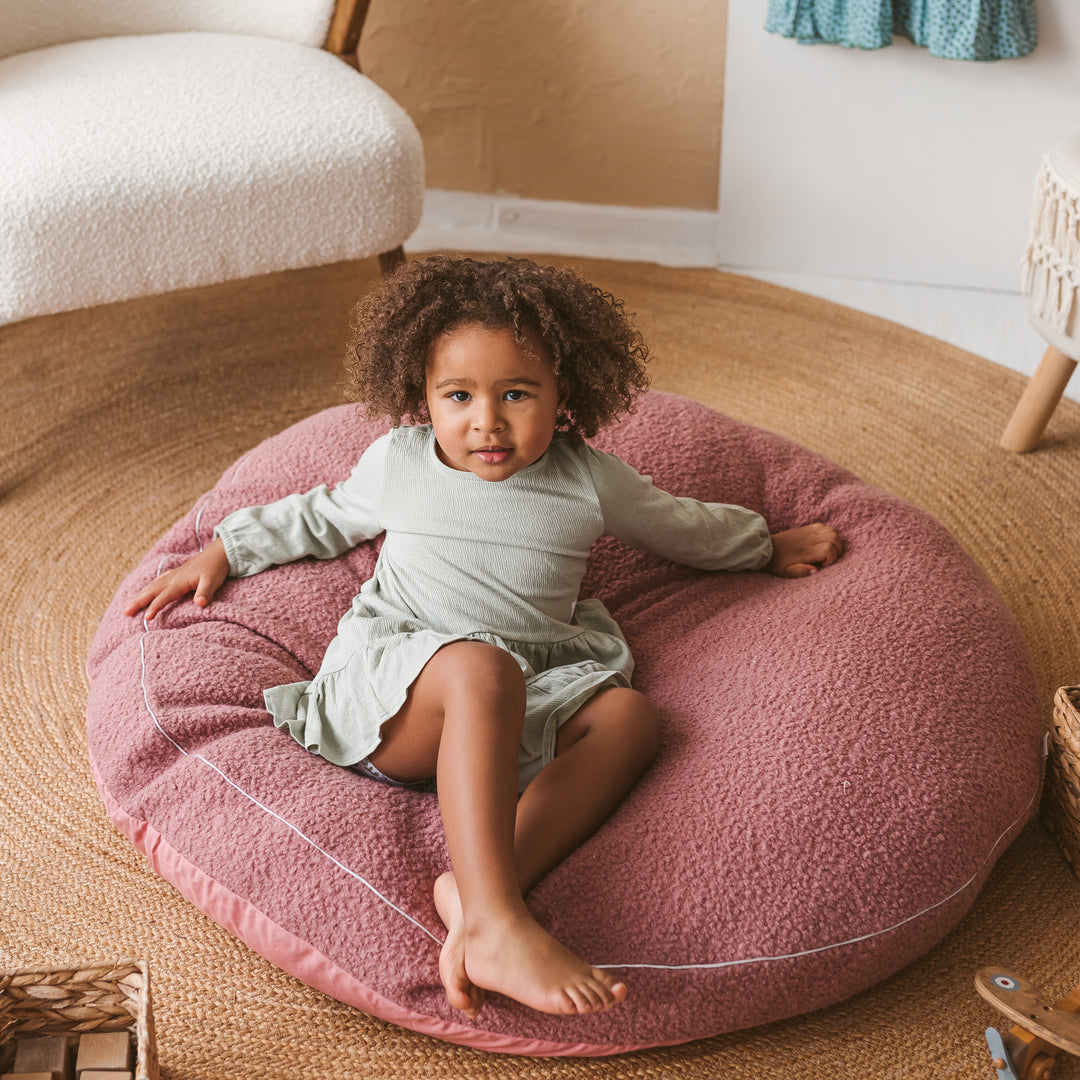 Overhead view of a child sitting cross-legged on the MINICAMP rose bouclé floor cushion, showing the textured fabric